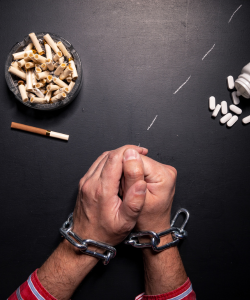 Symbolická fotografia ilustrujúca závislosť – ruky v reťaziach obklopené cigaretami a tabletkami, znázorňujúce silu návykov a zlozvykov. Tento obraz vyjadruje bezmocnosť a otroctvo spôsobené závislosťami, ako je fajčenie a užívanie látok.