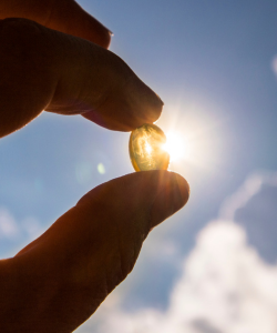 Kapsula vitamínu D držiaca v ruke na pozadí slnečného svetla, symbolizujúca význam vitamínu D pre vstrebávanie vápnika a zdravie kostí.